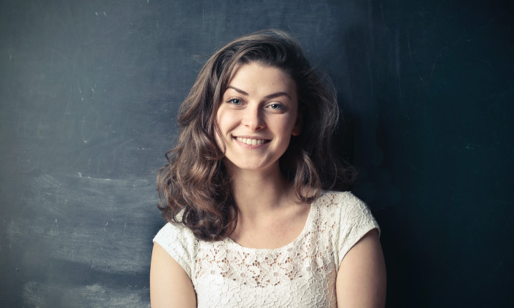 Brunette woman wearing a cream blouse smiles while standing against a black chalkboard