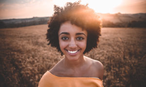 Curly-haired young woman smiles with teeth remineralized with fluoride wearing a yellow blouse in a wheat field