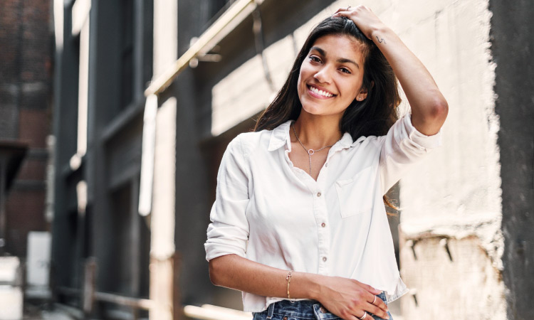 Brunette woman wearing a white blouse smiles with CEREC dental crowns in front of an old exterior wall of a building